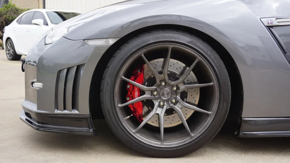 Close-up of a grey Nissan GT-R's front wheel, showing aftermarket multi-spoke wheels, red brake calipers, and carbon fiber aero.