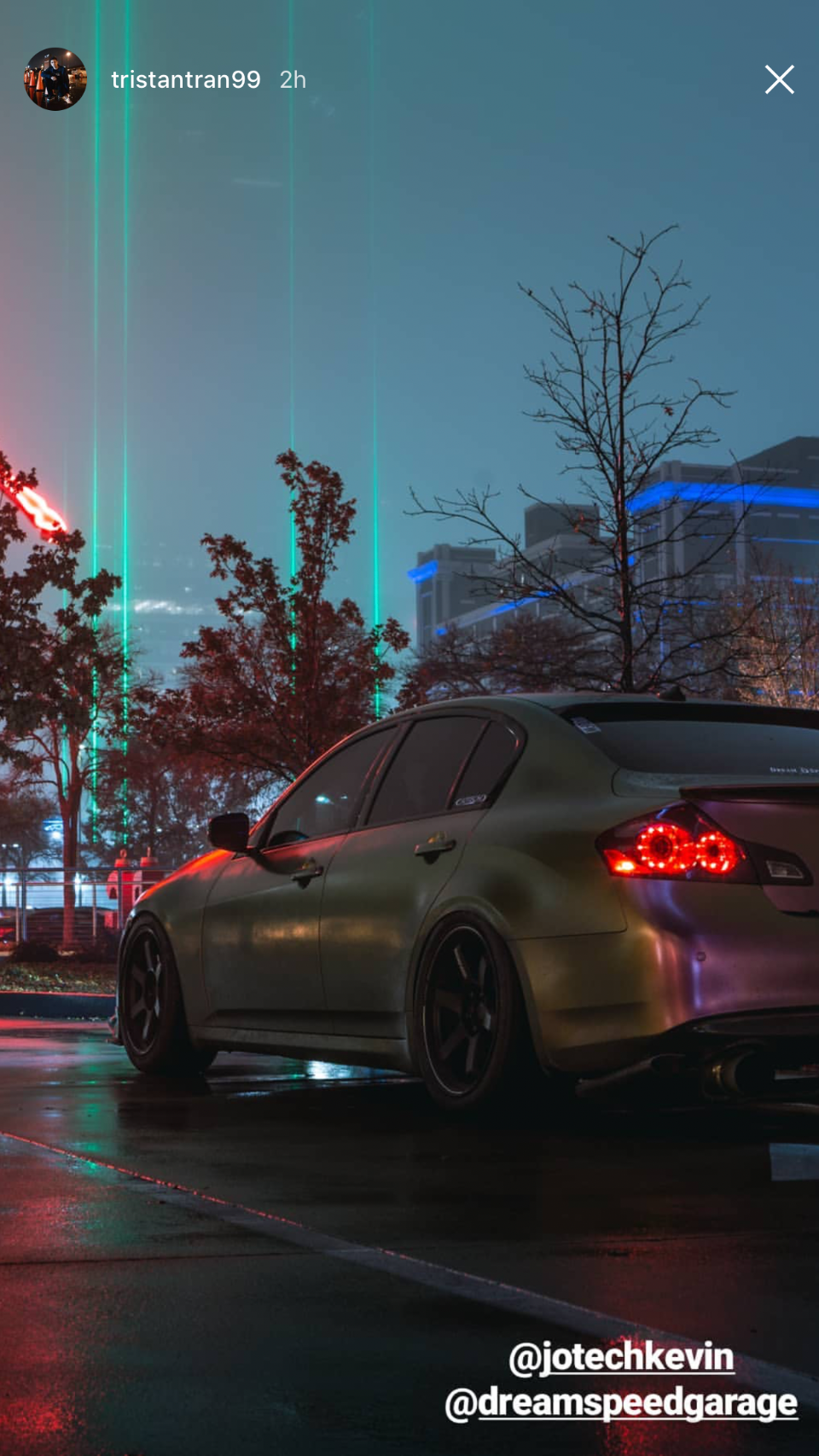 A modified Infiniti G37 sedan with aftermarket wheels and exhaust parked on a wet city street at night, illuminated by neon lights.