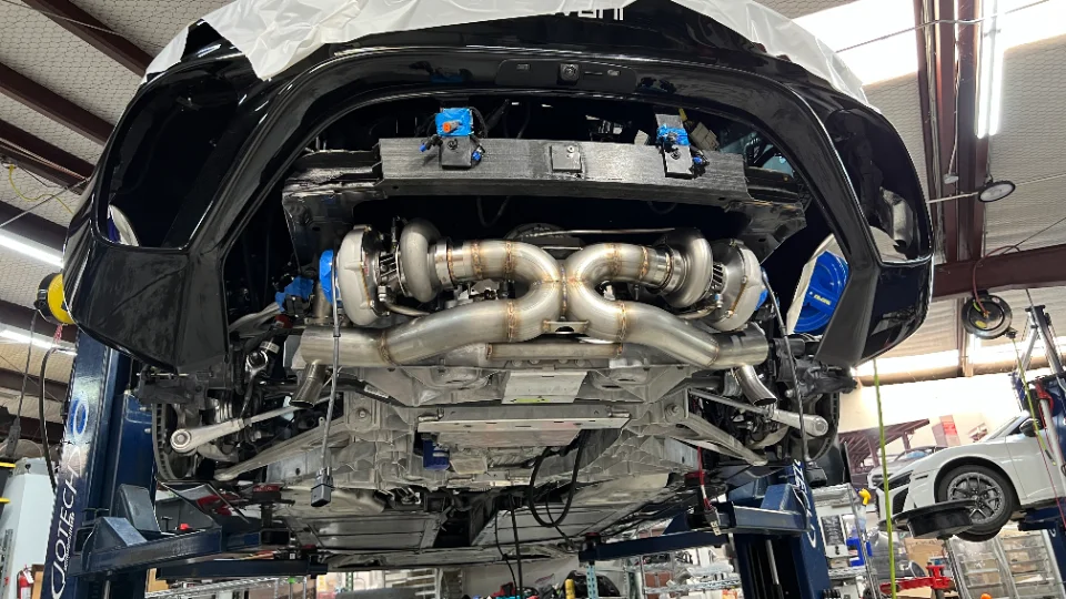 A black C8 Corvette on a lift in a performance shop, showcasing a custom twin-turbo system and exhaust fabrication.