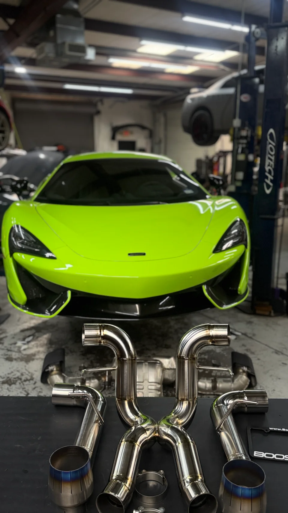 A bright green McLaren sports car on a lift in a shop, with a polished stainless steel aftermarket exhaust system laid out in the foreground.