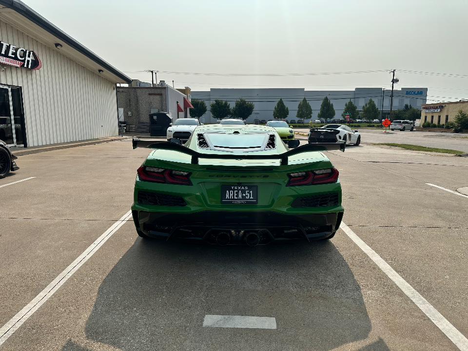 Rear view of a green Corvette C8 with a large aftermarket rear wing and custom exhaust parked in a lot outside a building.