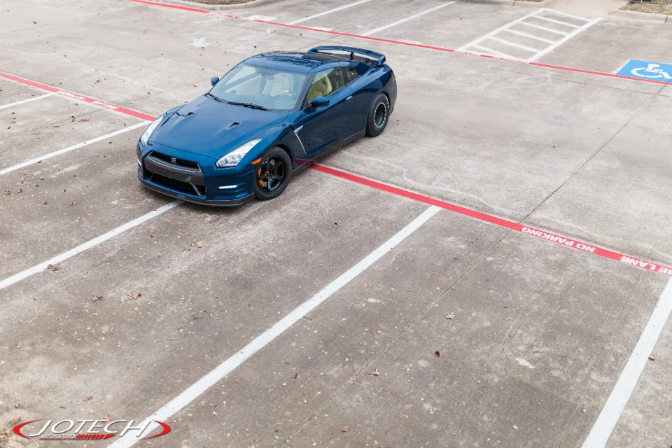 A blue Nissan GT-R R35 with aftermarket wheels and a spoiler parked in a concrete parking lot from an overhead view.
