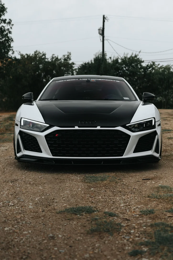 Front view of a white Audi R8 with a black carbon fiber hood and front splitter, parked on a dirt road.