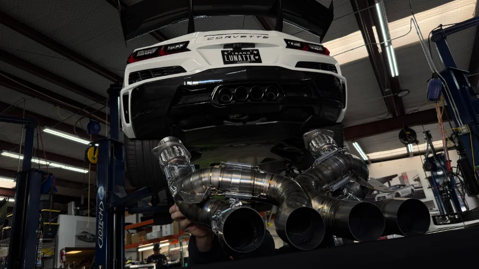 A white Corvette on a lift in a performance shop with a new aftermarket exhaust system being installed.