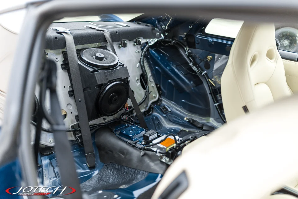 Interior view of a car undergoing modification, with rear seats removed exposing wiring harnesses, speakers, and chassis.