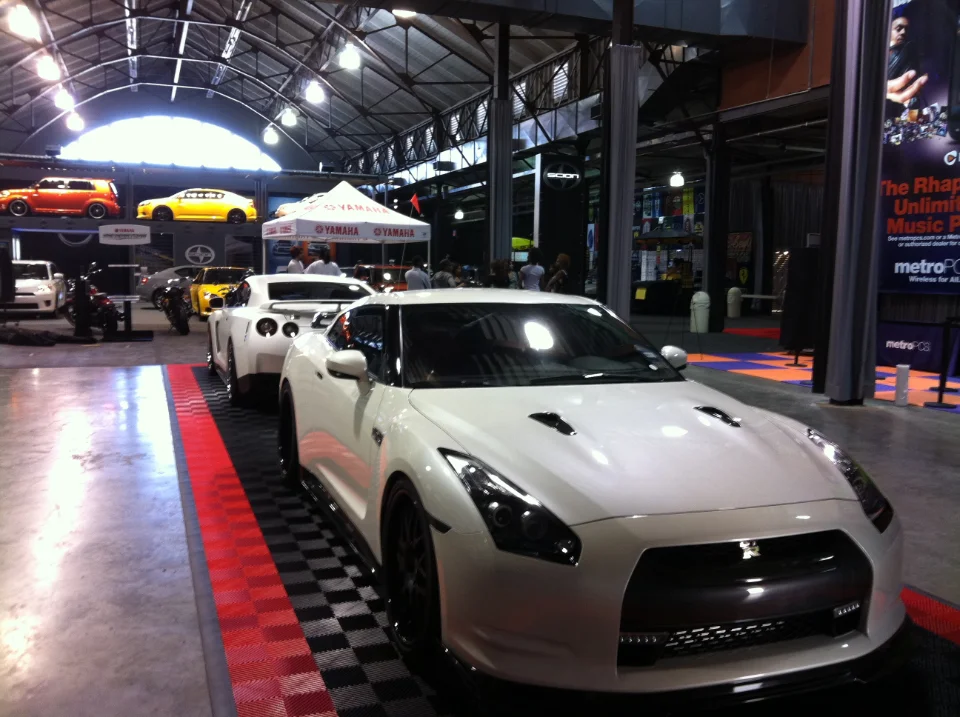 White Nissan GT-Rs on display at an indoor car show event with other vehicles and vendor booths in the background.