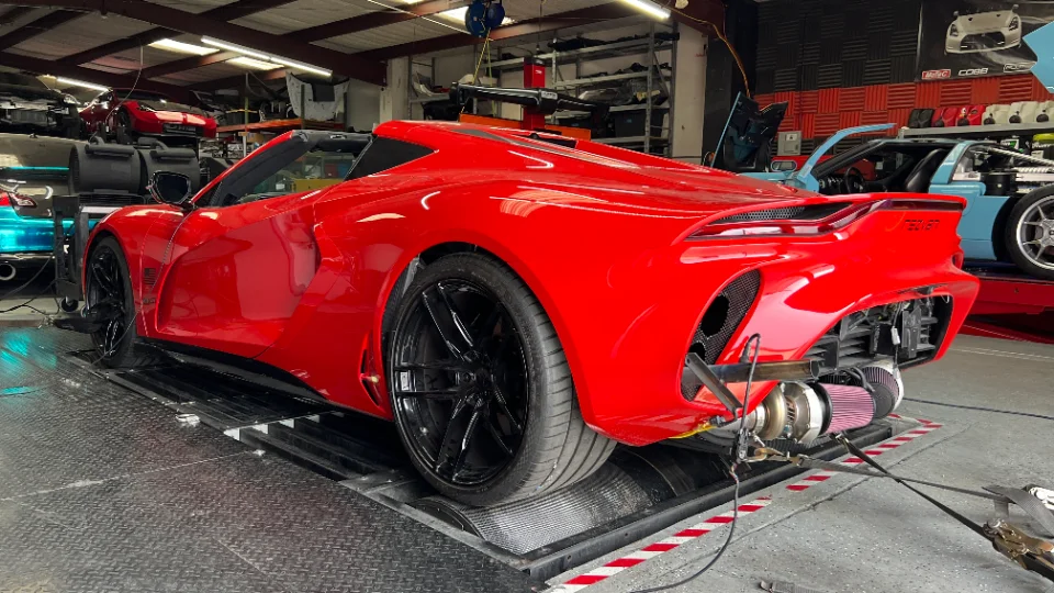 A red custom supercar on a dynamometer in a performance shop, with its rear bumper removed revealing a large turbocharger and air filters.