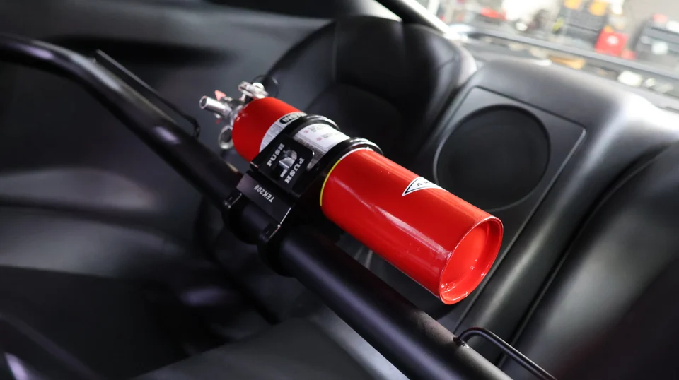A red fire extinguisher mounted on a black roll bar inside a performance car's interior, emphasizing safety equipment.