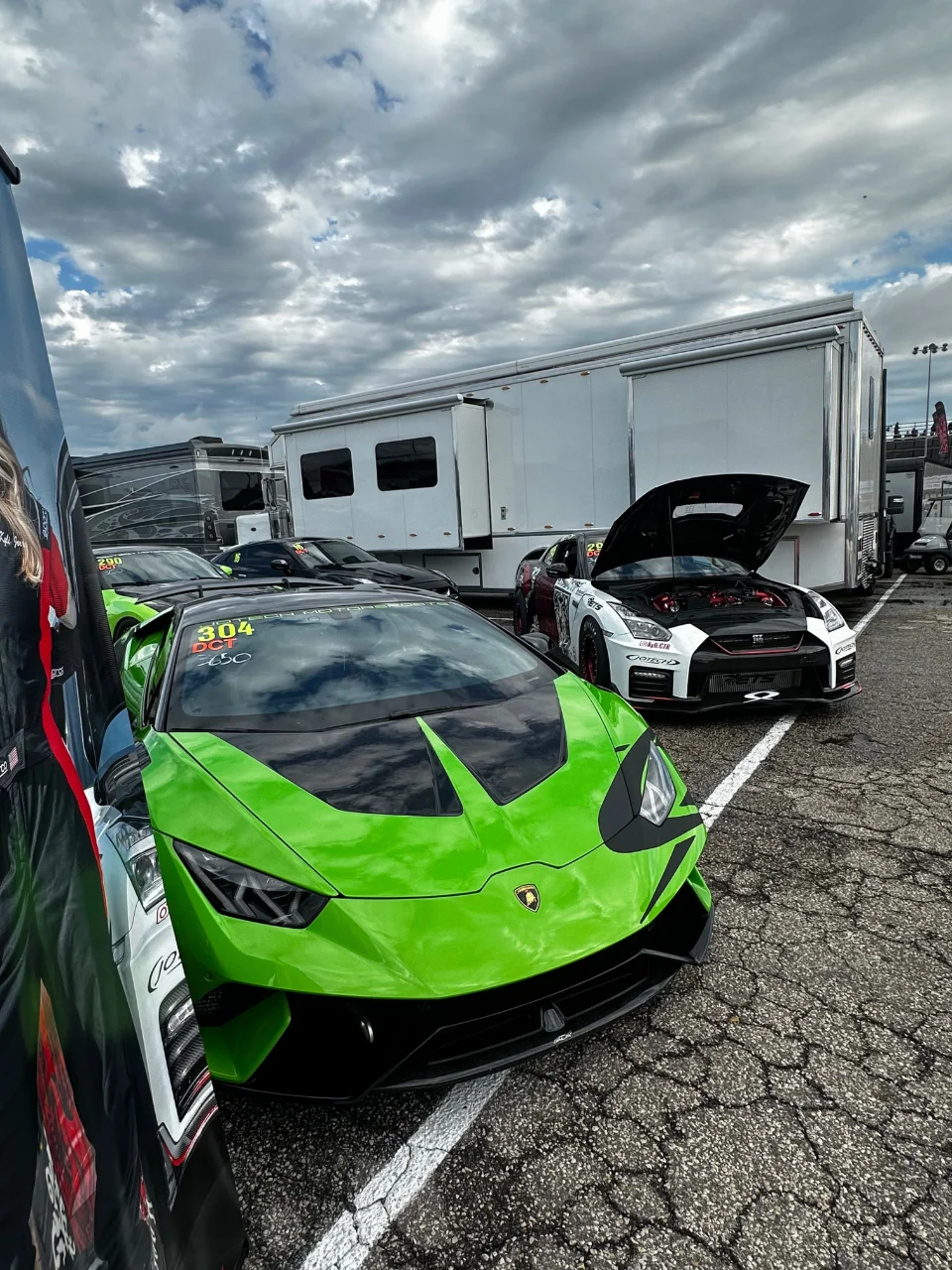 A bright green Lamborghini Huracan and a white Nissan GT-R with its hood open parked in a track paddock area with large trailers.