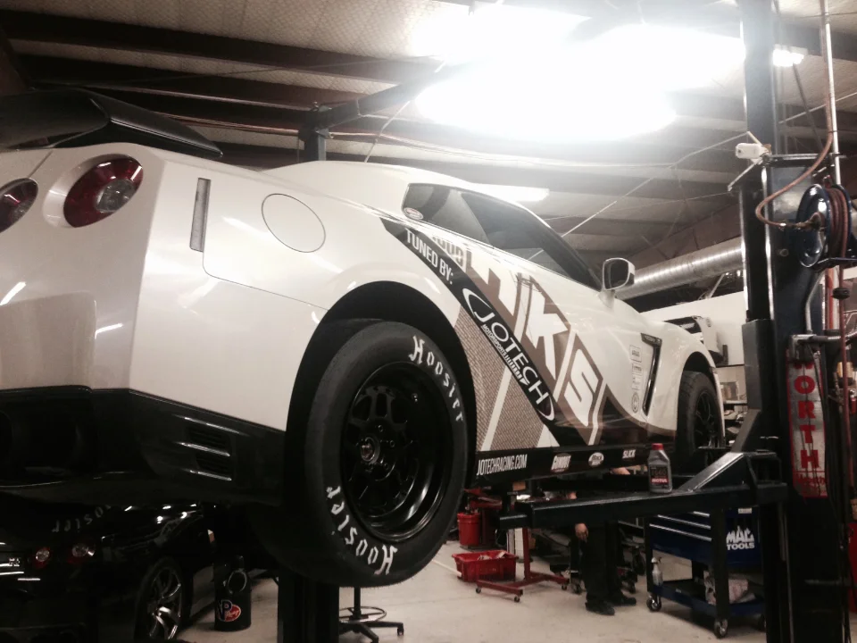White Nissan GT-R race car with HKS Jotech livery on a lift in a performance tuning shop, featuring Hoosier drag slicks.