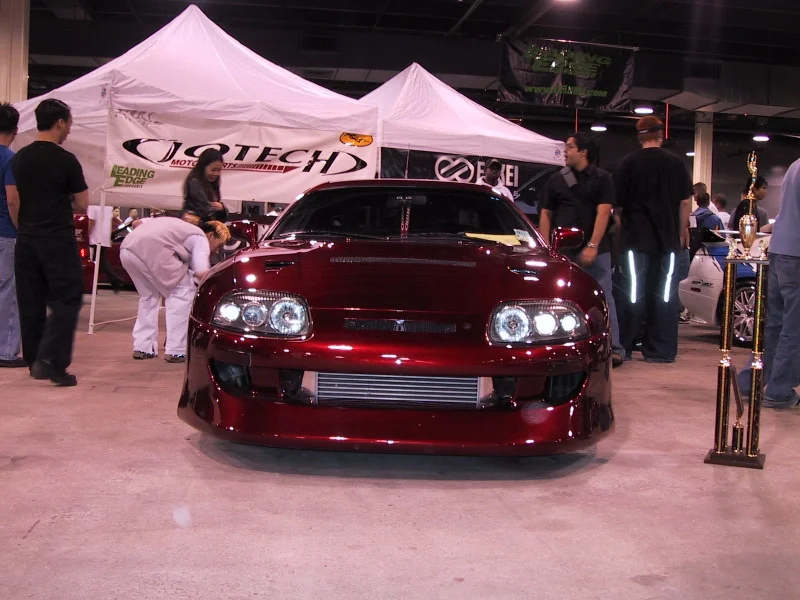 A custom red Toyota Supra on display at a car show, with an intercooler visible and a trophy next to it.
