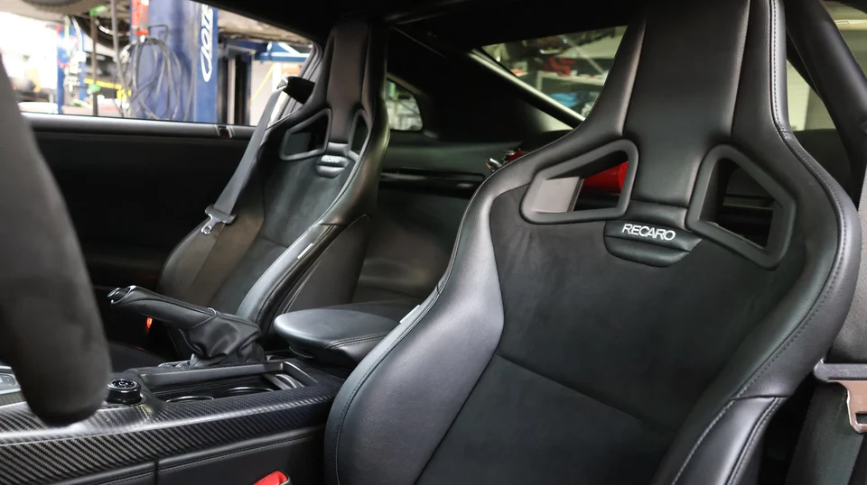 Interior view of a performance car, likely a GT-R, featuring Recaro bucket seats and carbon fiber trim, with a shop environment visible in the background.