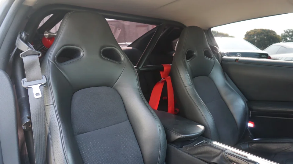 Interior view of a Nissan GT-R featuring black racing seats, red racing harnesses, a black roll cage, and a fire extinguisher.