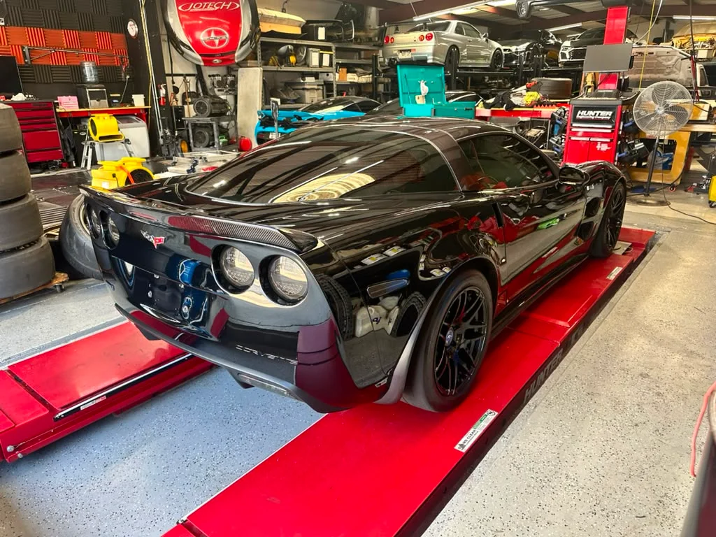 A black C6 Corvette on an alignment rack inside a performance tuning shop with other cars and equipment in the background.