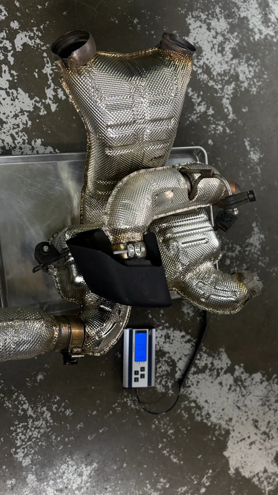 A heat-shielded exhaust component, possibly a downpipe, being weighed on a digital scale in a shop.