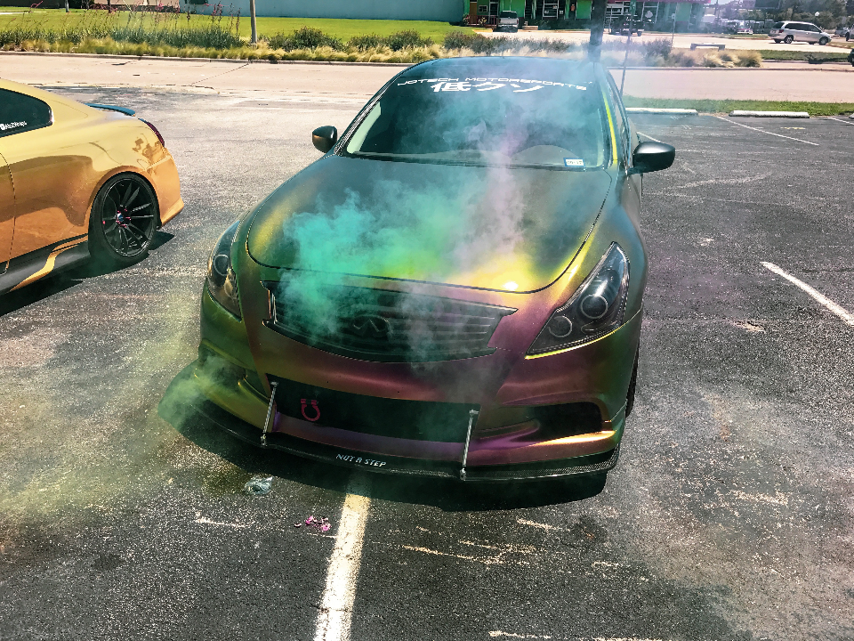 An Infiniti G37 with a chameleon wrap and front splitter, surrounded by colored smoke in a parking lot.
