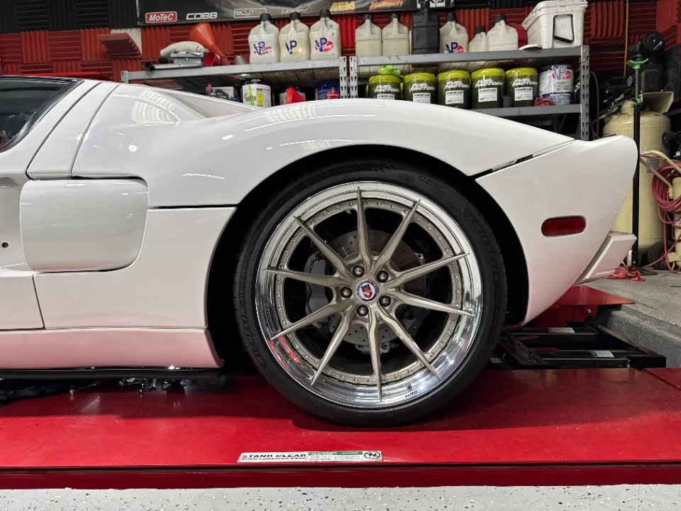 A white Ford GT on a red lift in a performance shop, showcasing custom HRE wheels and rear suspension.