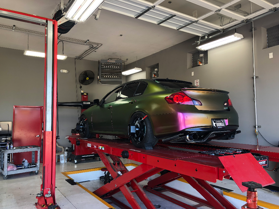 An Infiniti G37 with a chameleon wrap and aftermarket exhaust system on a red scissor lift in a performance tuning shop.