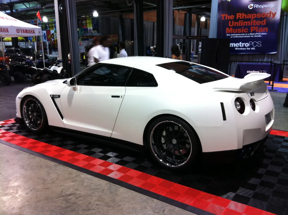 White Nissan GT-R with aftermarket wheels and a rear wing displayed on a checkered floor at an indoor event.