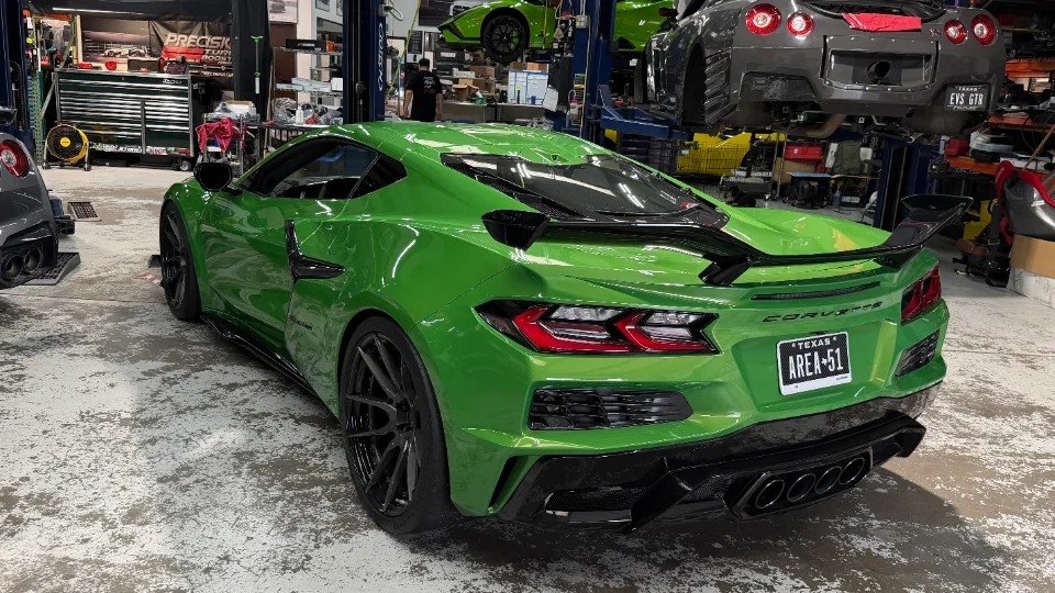 A bright green Chevrolet Corvette C8 with aftermarket wheels and a large rear wing parked in a performance tuning shop.