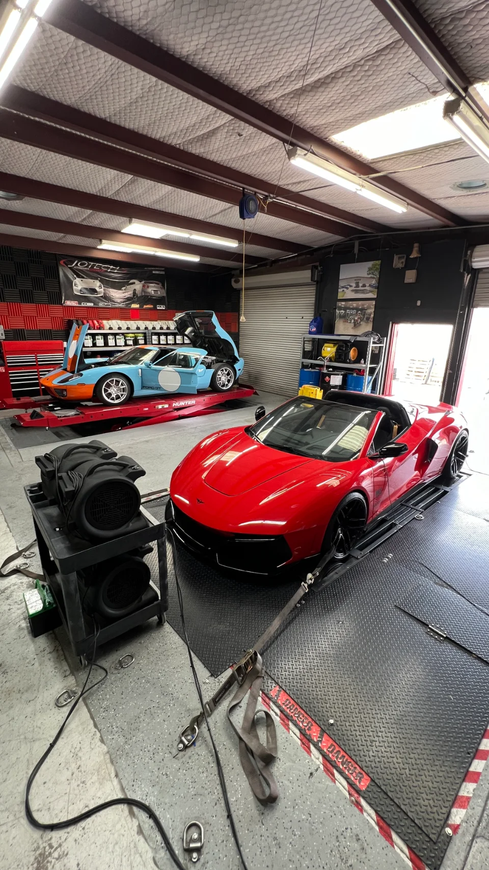 A red McLaren supercar strapped to a dyno with a blue and orange Ford GT on a car lift in a performance tuning shop.