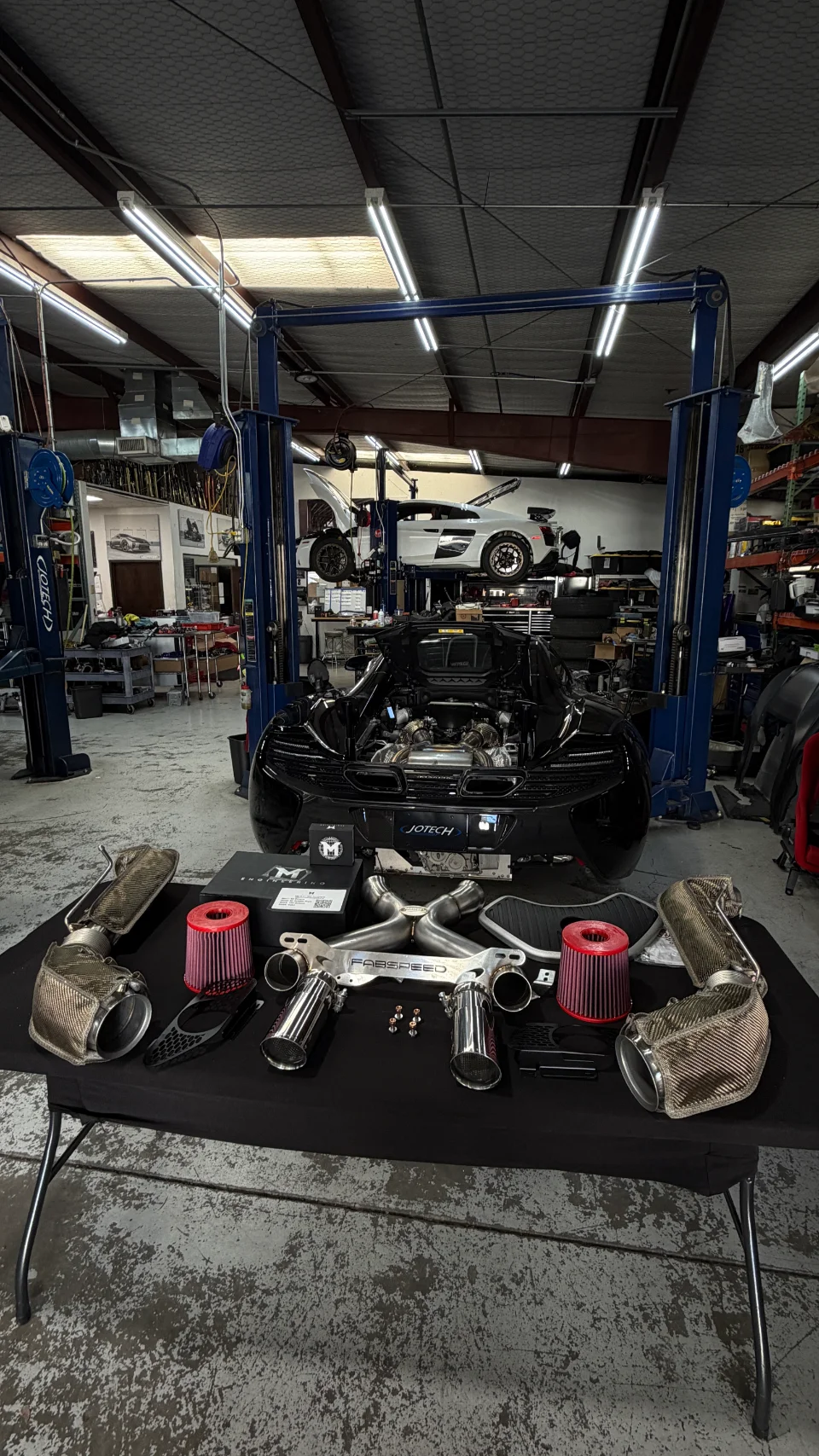 McLaren with its engine bay open in a performance shop, surrounded by aftermarket exhaust components and air filters on a table.