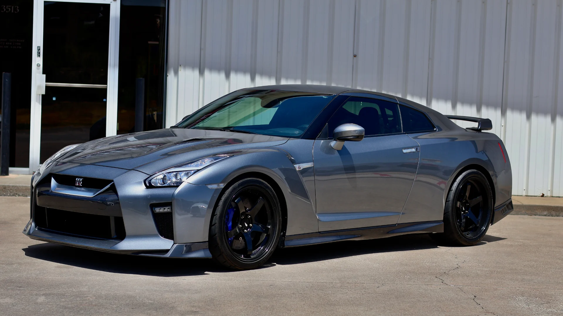 A sleek grey Nissan GT-R with black aftermarket wheels parked outside a modern building on a sunny day.