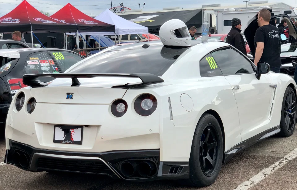A white Nissan GT-R with race number 181 and a helmet on the roof, parked at a drag strip with tents in the background.