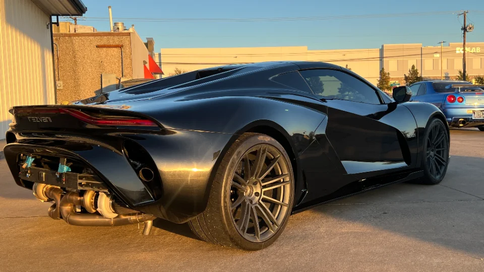 A black Rezvani supercar with exposed turbochargers and custom exhaust, parked outdoors with a blue Nissan GT-R in the background.