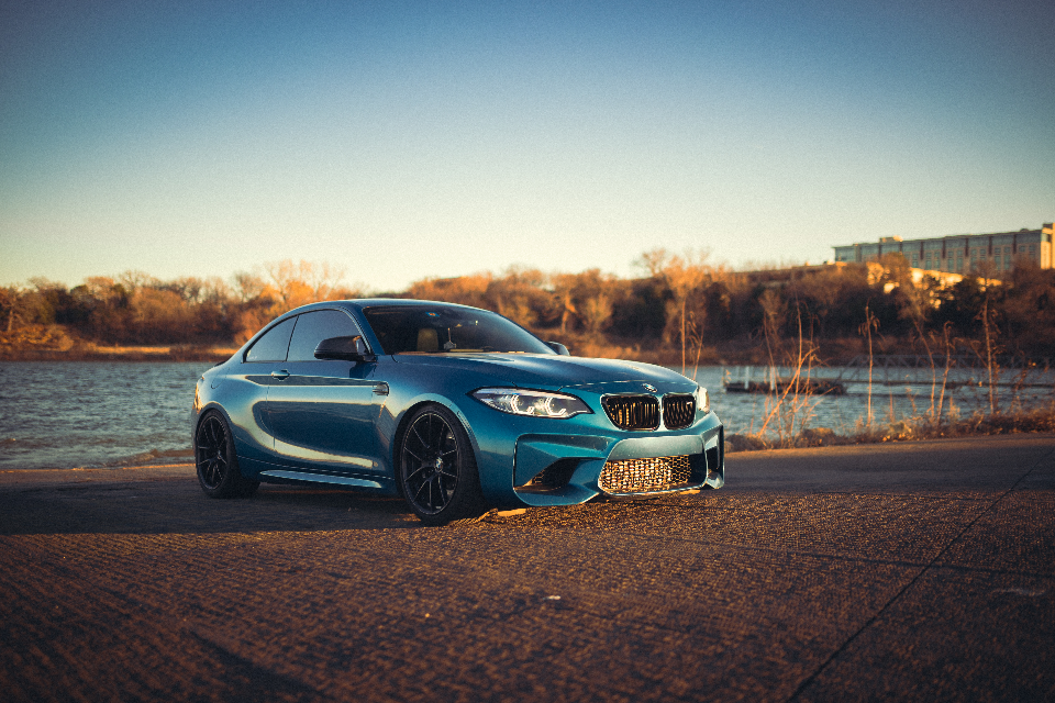 A blue BMW M2 performance car parked by a lake at sunset, showcasing its sleek design and black wheels.
