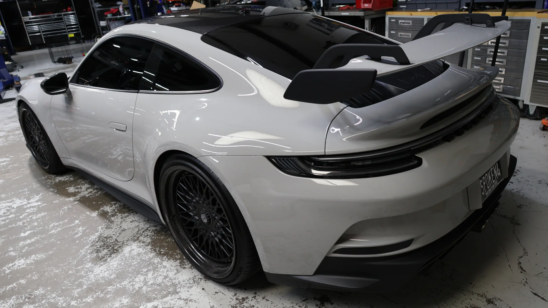A white Porsche 911 with aftermarket black multi-spoke wheels and a large rear wing parked in a performance shop.