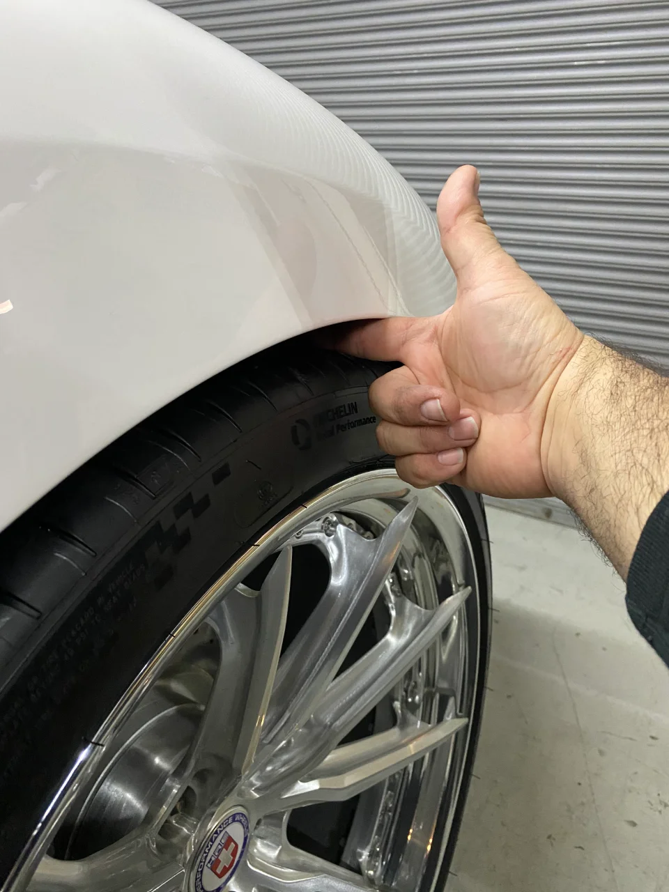 Close-up of a white car's front wheel and fender, showing tire fitment with a hand for scale. Features HRE Performance Wheels and Michelin tires.