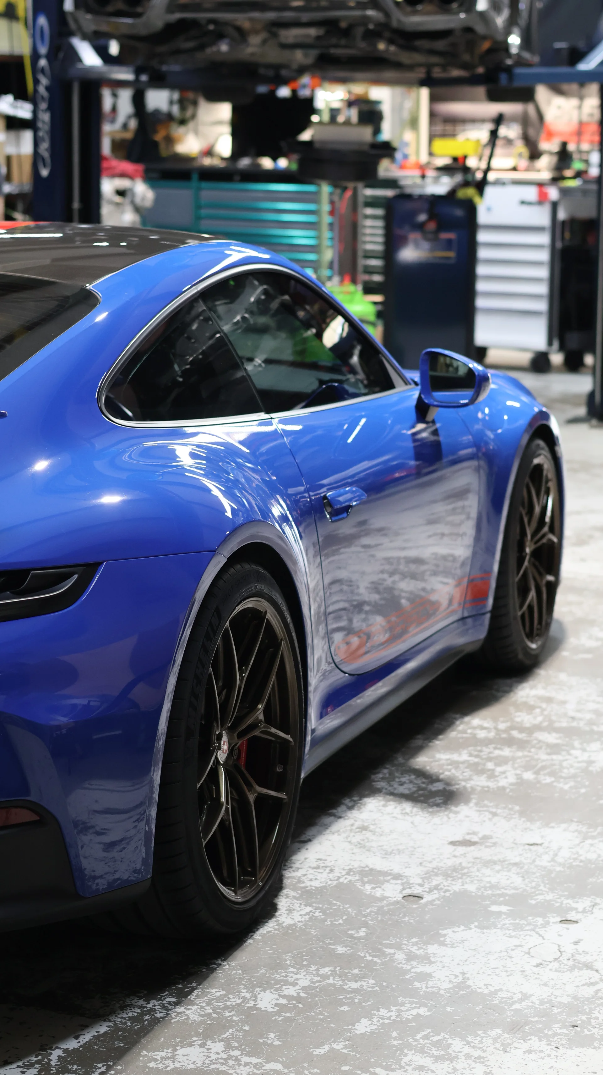 A blue Porsche sports car parked in a clean automotive shop with a car lift and toolboxes in the background.