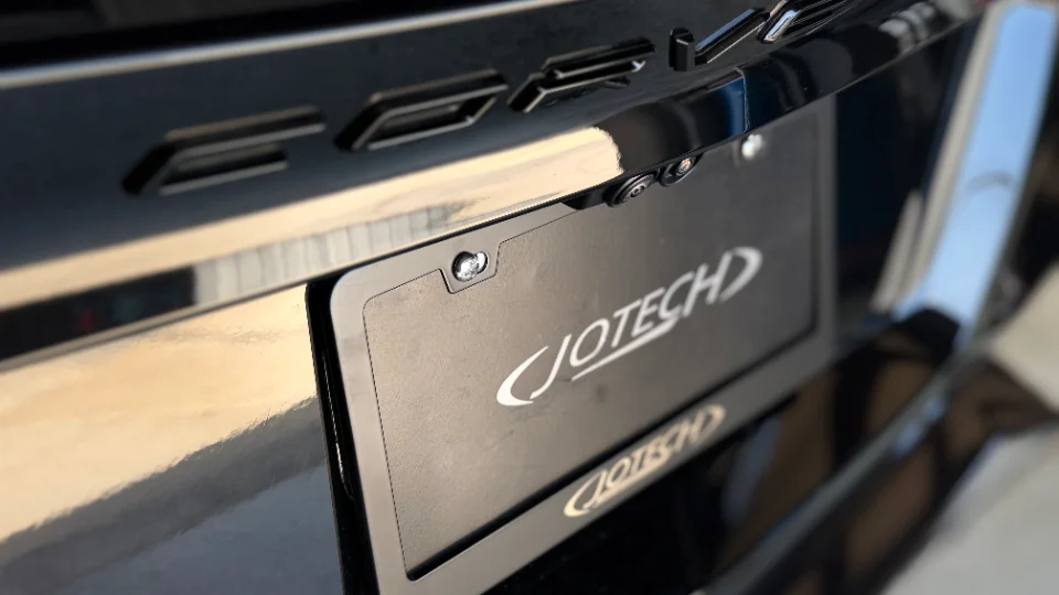 A close-up detail shot of a black car's rear bumper featuring a Jotech branded license plate frame and backup camera.
