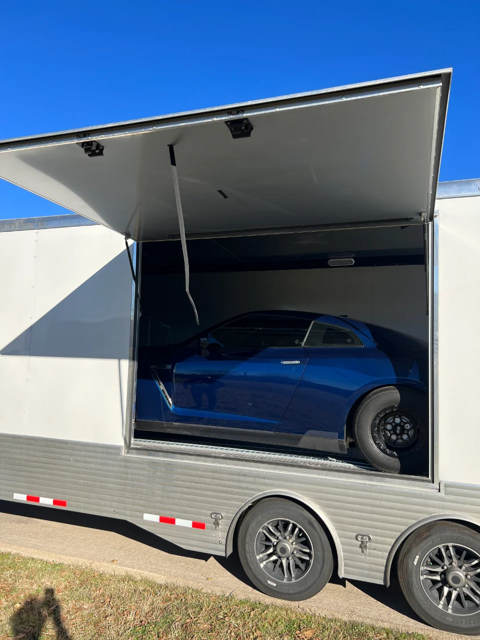A blue Nissan GT-R with drag racing wheels loaded inside an open enclosed car trailer under a clear blue sky.