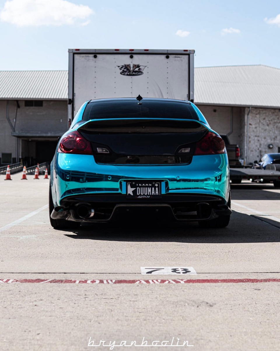 Rear view of a teal chrome-wrapped Infiniti G37 coupe with a custom exhaust and spoiler, parked outdoors.