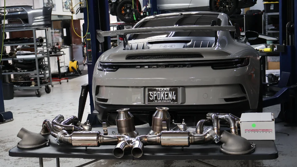 A grey Porsche 911 GT3 on a lift in a performance shop with a Fabspeed exhaust system laid out in the foreground.