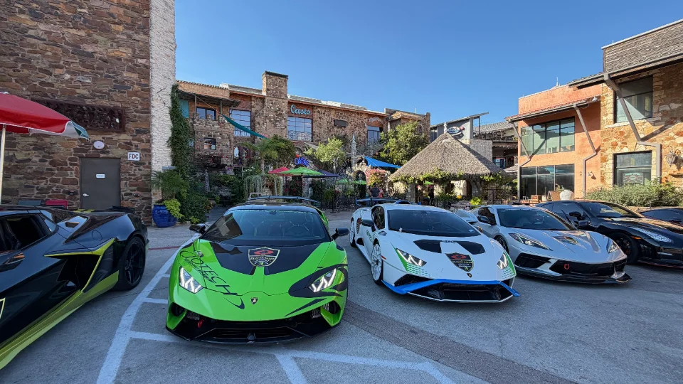 A group of high-performance cars, including a green and a white Lamborghini Huracan and a silver Corvette, parked outside a building.