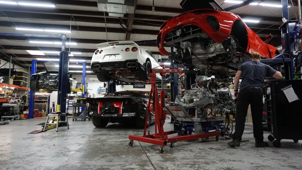 A mechanic works on a red sports car with its engine removed, while a white Nissan GT-R is on a lift in a busy performance shop.