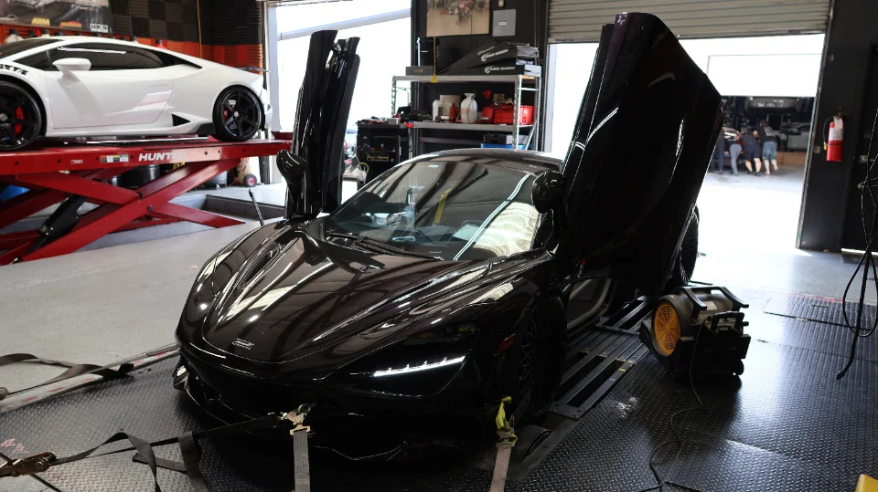 Black McLaren 720S strapped to a dyno with its doors open, and a white Lamborghini Huracan on a lift in the background at a performance shop.