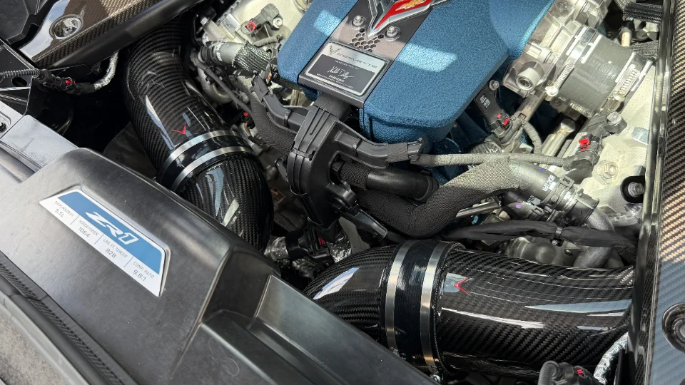 A close-up of a Corvette ZR1 engine bay featuring carbon fiber intake tubes and a blue air filter box.
