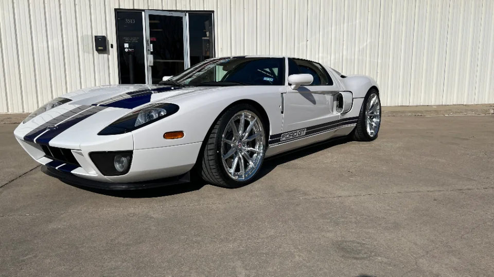 A white Ford GT with blue racing stripes and custom chrome wheels parked in front of a building on a sunny day.