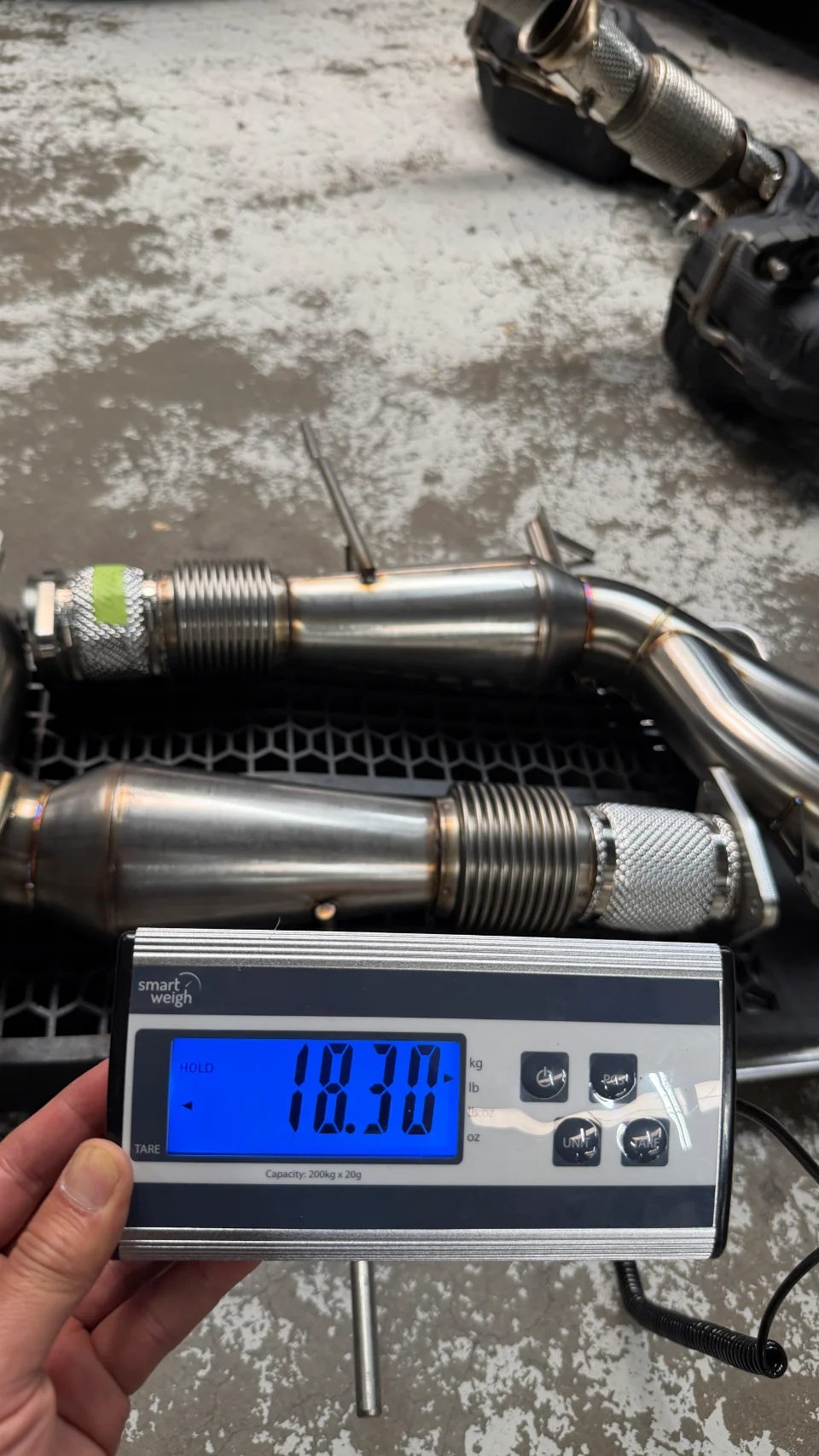 Stainless steel exhaust downpipes being weighed on a digital scale, showing a weight of 18.30 lbs.