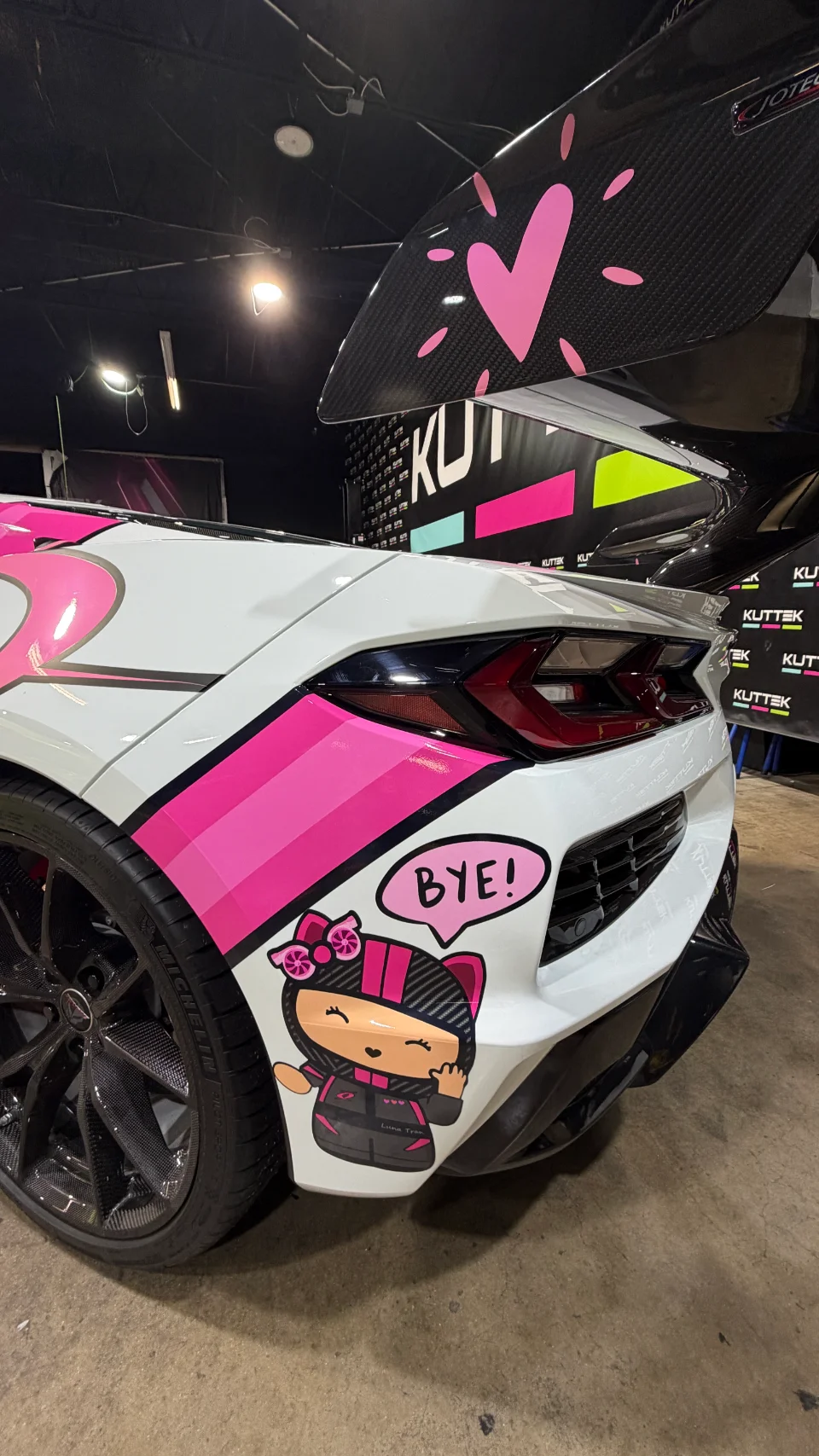 Rear view of a white C8 Corvette with a custom pink and black livery, featuring a carbon fiber rear wing and 'BYE!' graphic.