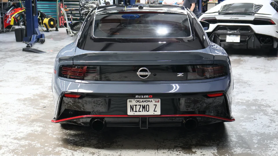 Rear view of a grey Nissan Z Nismo with custom exhaust in a performance shop, with a white Lamborghini in the background.