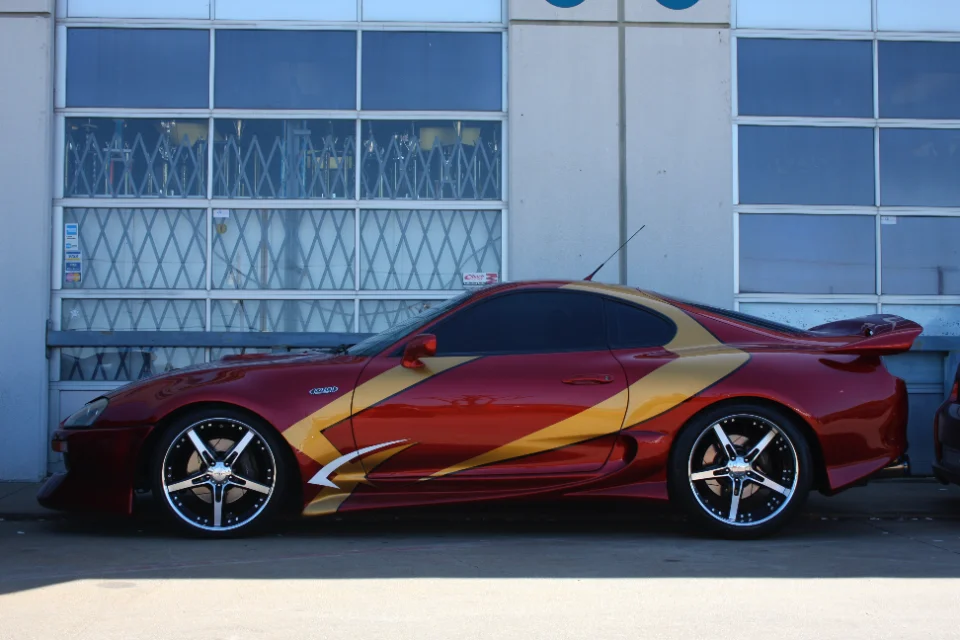 A red Toyota Supra with custom gold and white graphics and aftermarket chrome wheels parked outside a building.