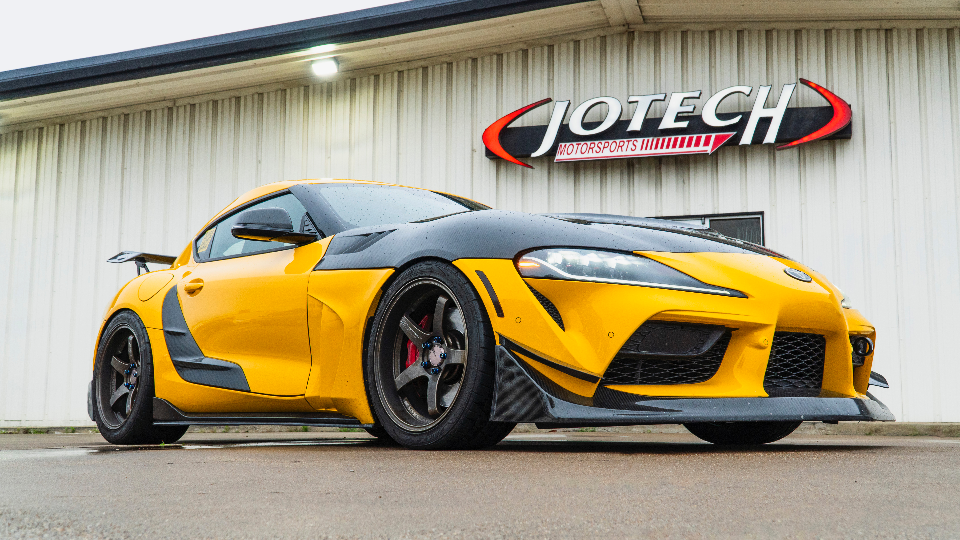 Yellow Toyota Supra with black carbon fiber hood and aero kit parked in front of Jotech Motorsports building.