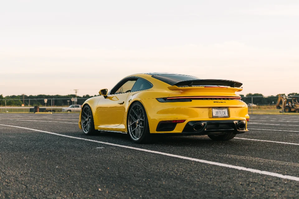 A vibrant yellow Porsche 911 Turbo S with aftermarket wheels and a prominent rear spoiler, parked on an asphalt lot at sunset.