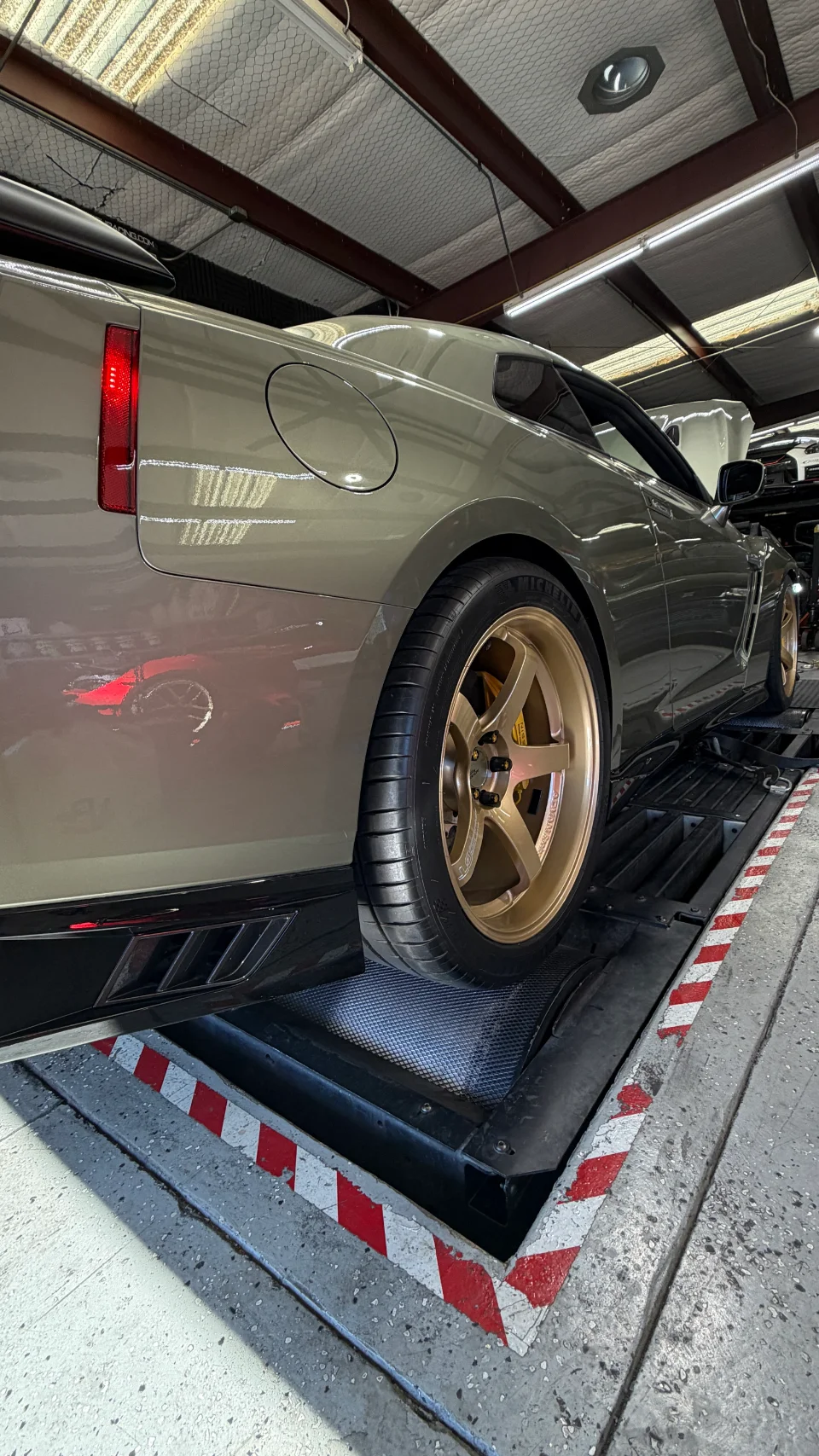 A Nissan GT-R in a matte olive green finish with gold aftermarket wheels strapped to a dynamometer at a performance shop.