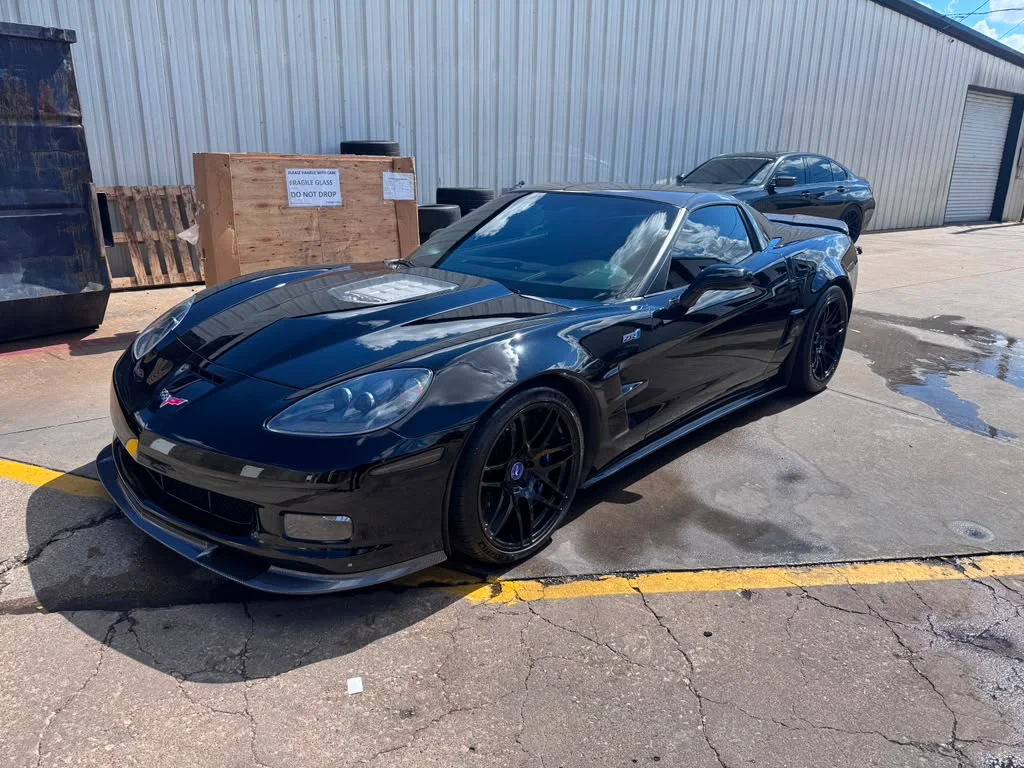 A black Chevrolet Corvette C6 Z06 with aftermarket wheels and a front splitter parked outside a shop.
